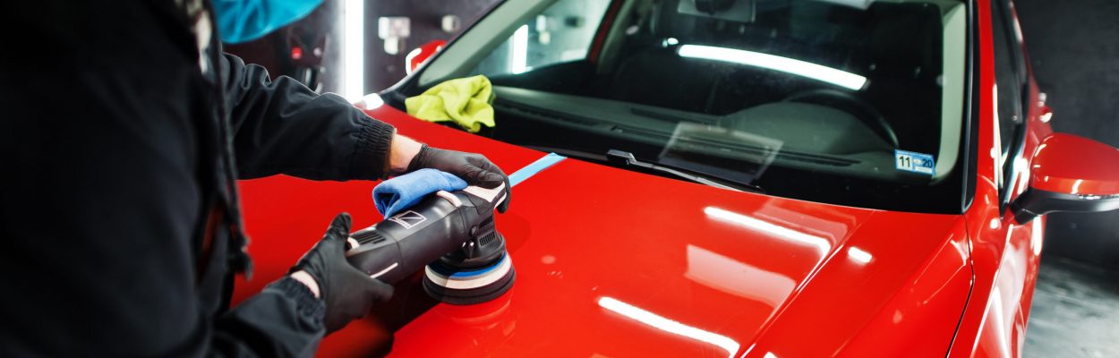 Car detailing concept. Man in face mask with orbital polisher in repair shop polishing orange suv car.