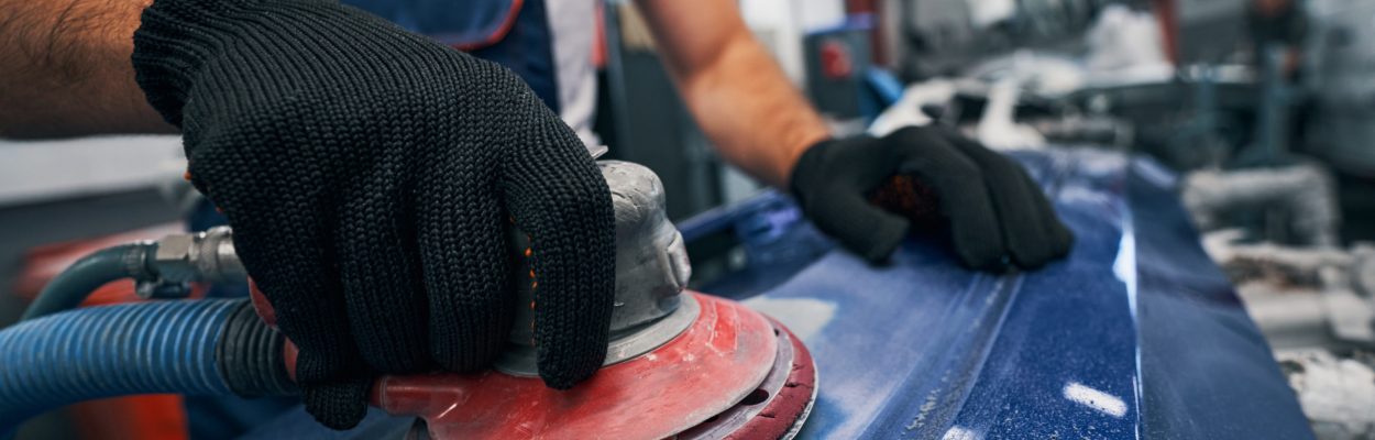 Automobile mechanic in black work gloves using grinder on blue metal surface of car spare part